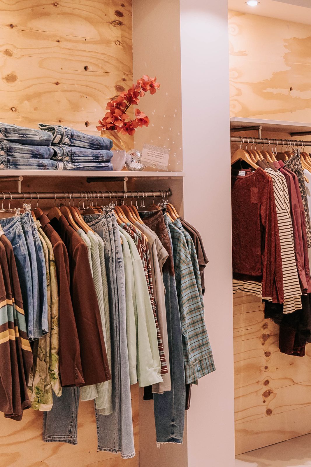 Rekken met blouses, overhemden en jeans tegen een houten wand in de winkel van KUIF