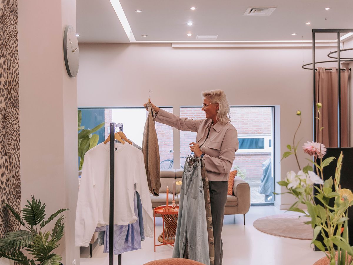 Ellie hangt in de lichte winkel van KUIF een jeans aan het rek, naast een vaas met bloemen