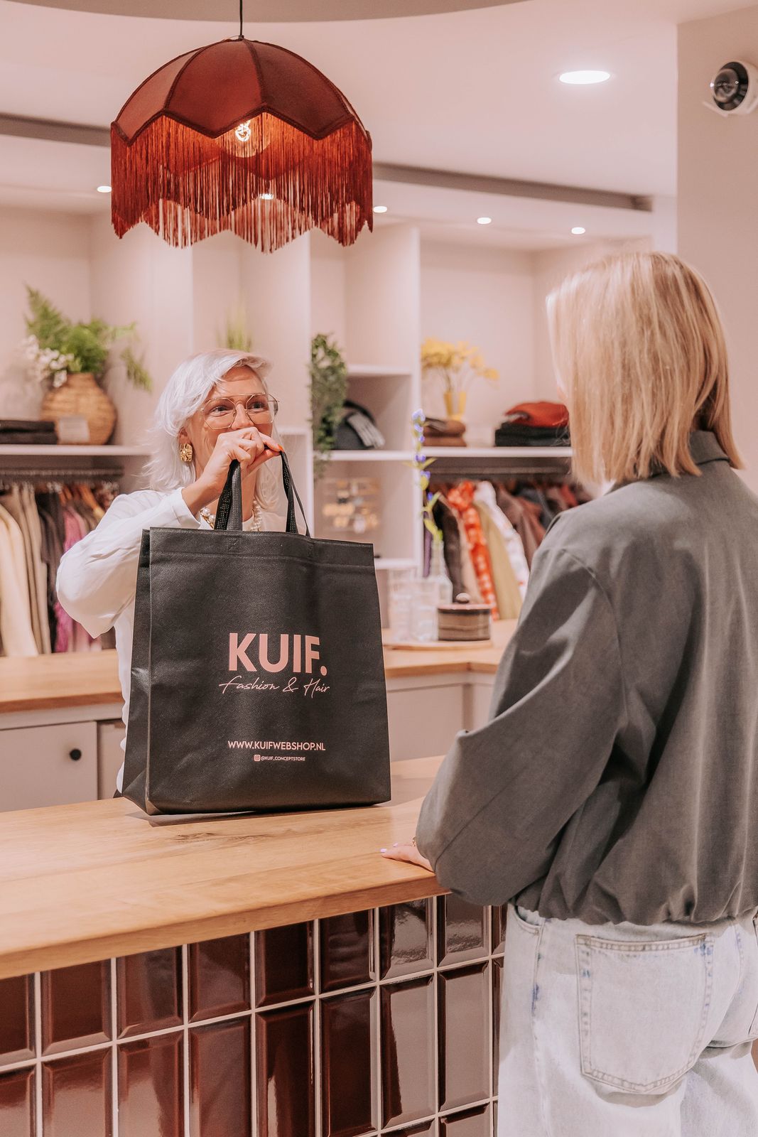 Ellie geeft onder de franjelamp een volle KUIF-tas aan een klant over de toonbank