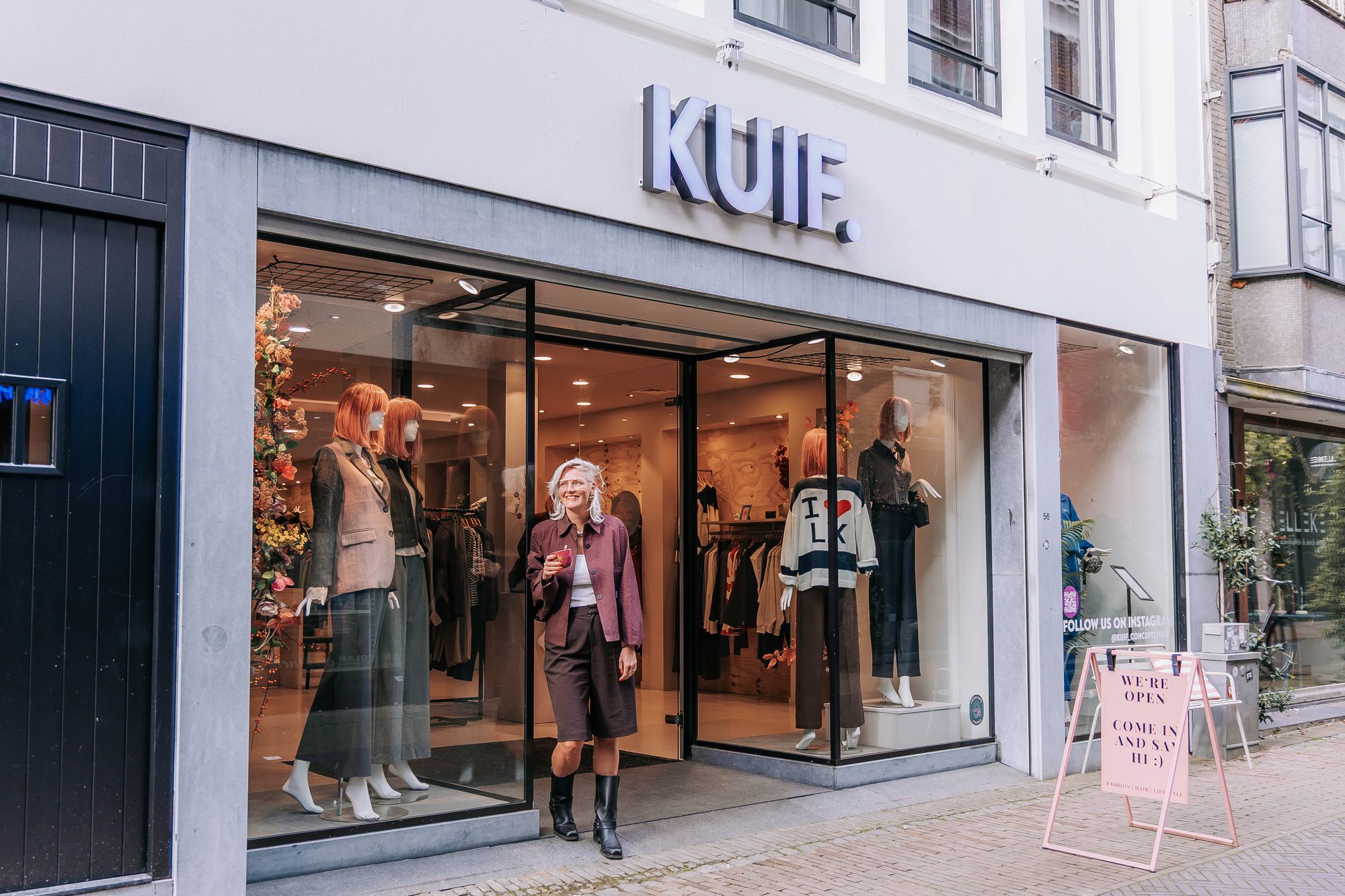 Ellie stapt met een kop koffie de open deur van KUIF uit, tussen etalages met paspoppen en onder het woordmerk op de gevel